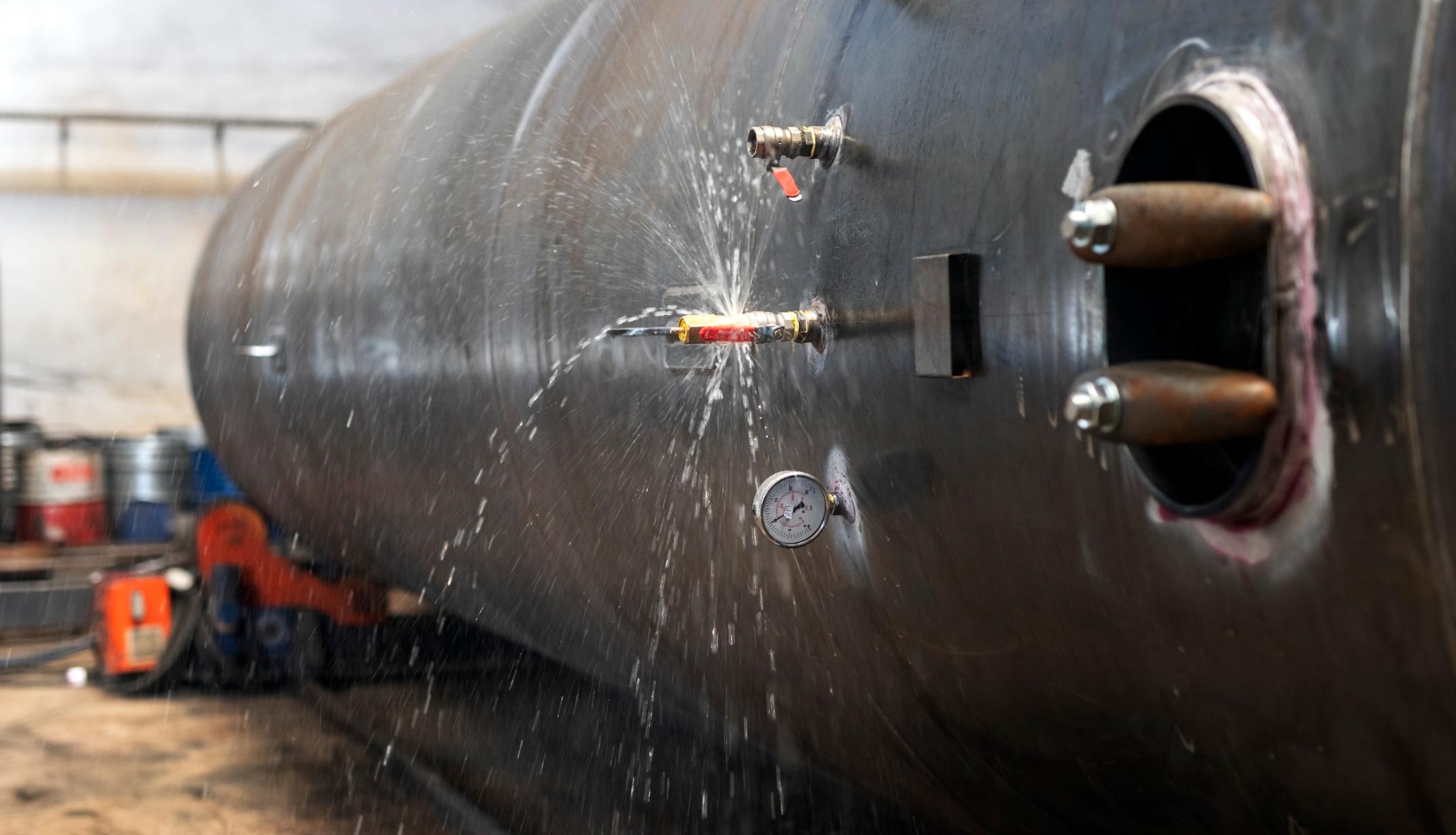 Water spraying from a leaking pipe connection on a large industrial metal tank in a workshop.