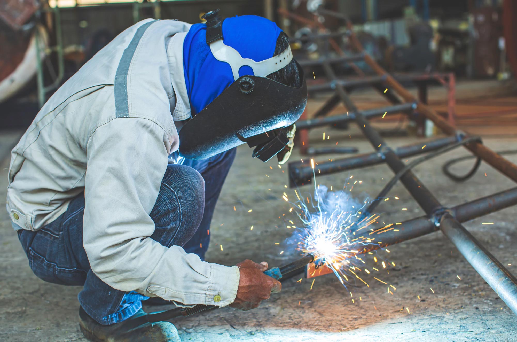A person in safety gear crouches and welds metal pipes, with bright sparks flying.