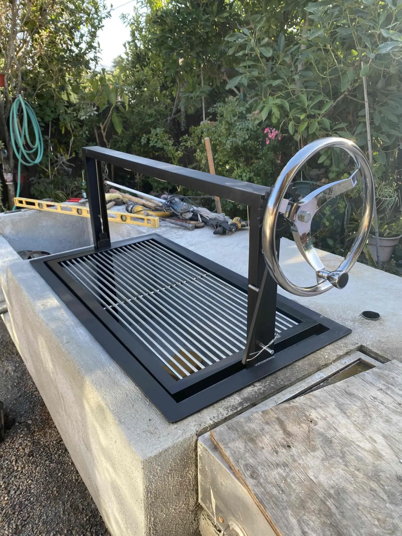 IMG_5071 Outdoor grill with a metal grate and a hand wheel for adjusting height, set into a concrete countertop.