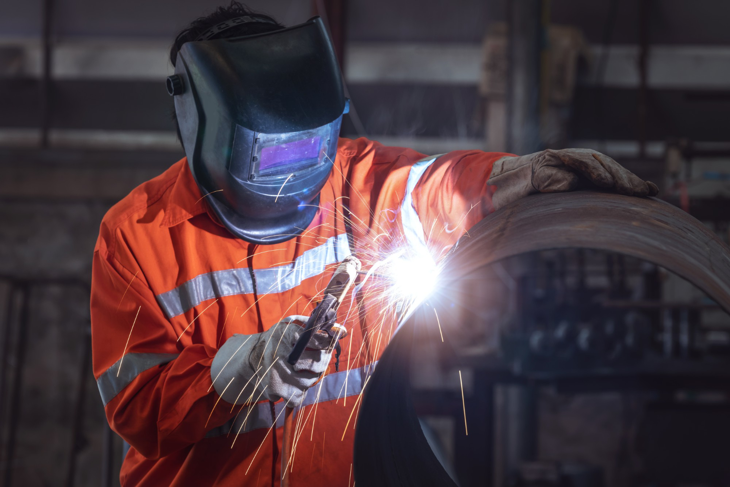 A worker in an orange suit and welding mask welds a large metal pipe, sparks flying as they ensure top welding quality.