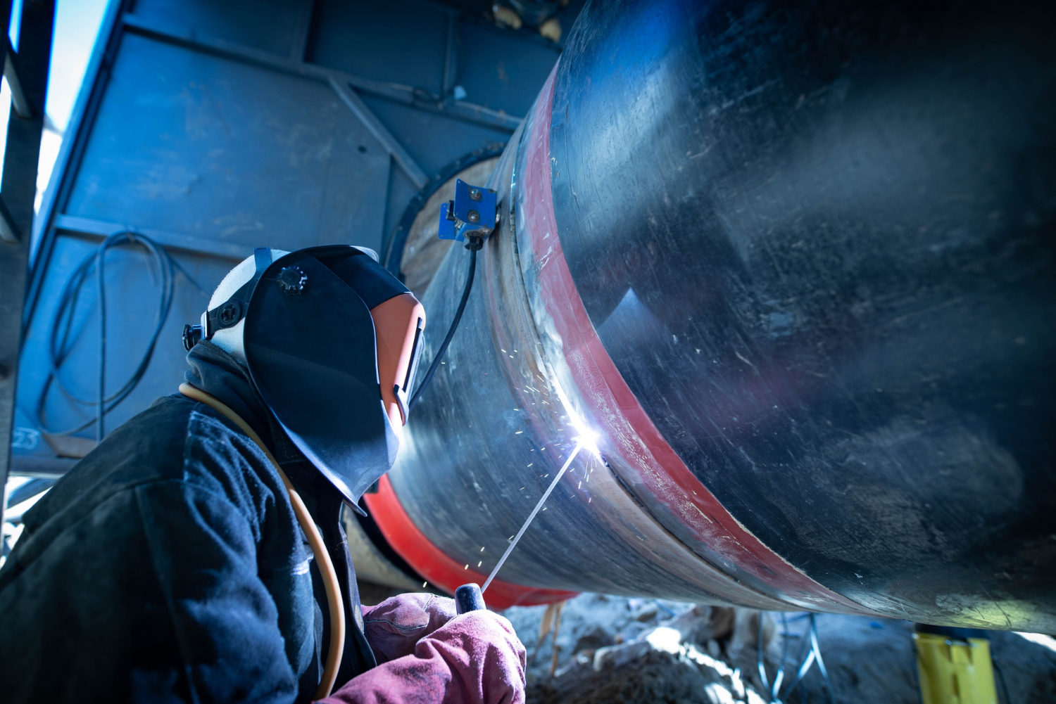 1573 A worker in protective gear welds a large metal pipe outdoors, creating bright sparks.