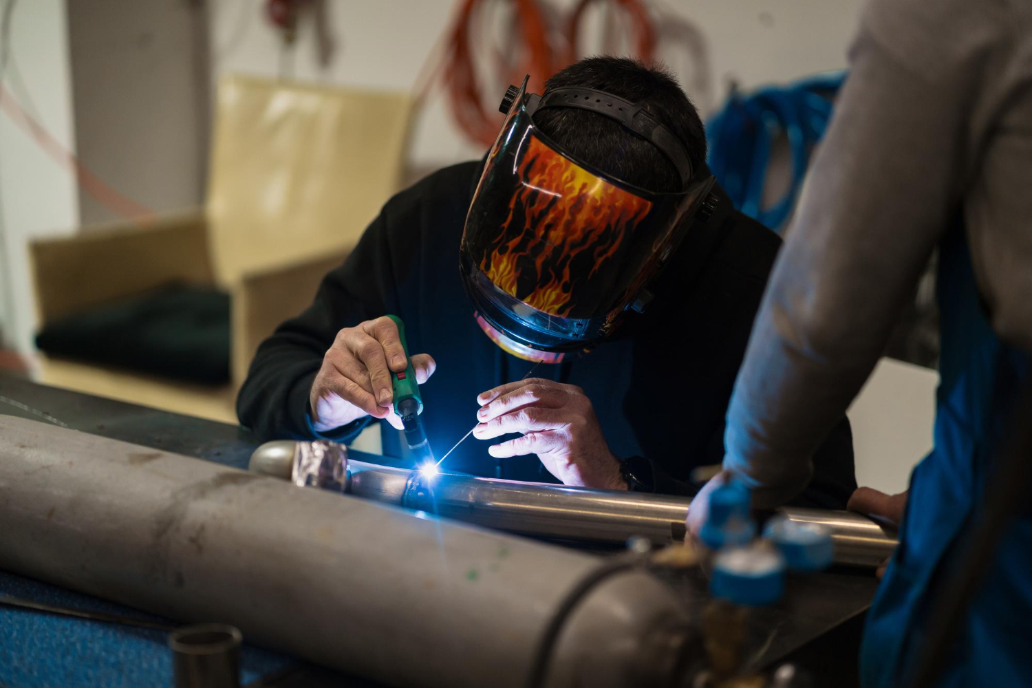 Person wearing a flame-patterned welding helmet welds metal pipes in a workshop, sparks visible.