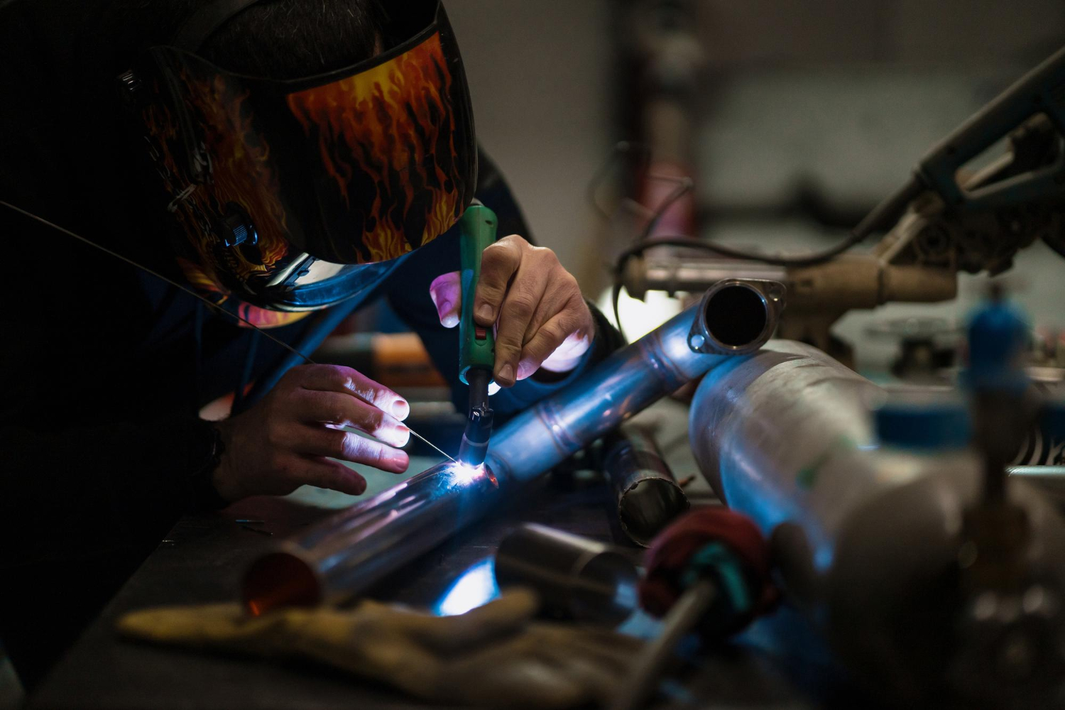 A person welding metal pipes while wearing a protective mask, surrounded by tools and equipment.