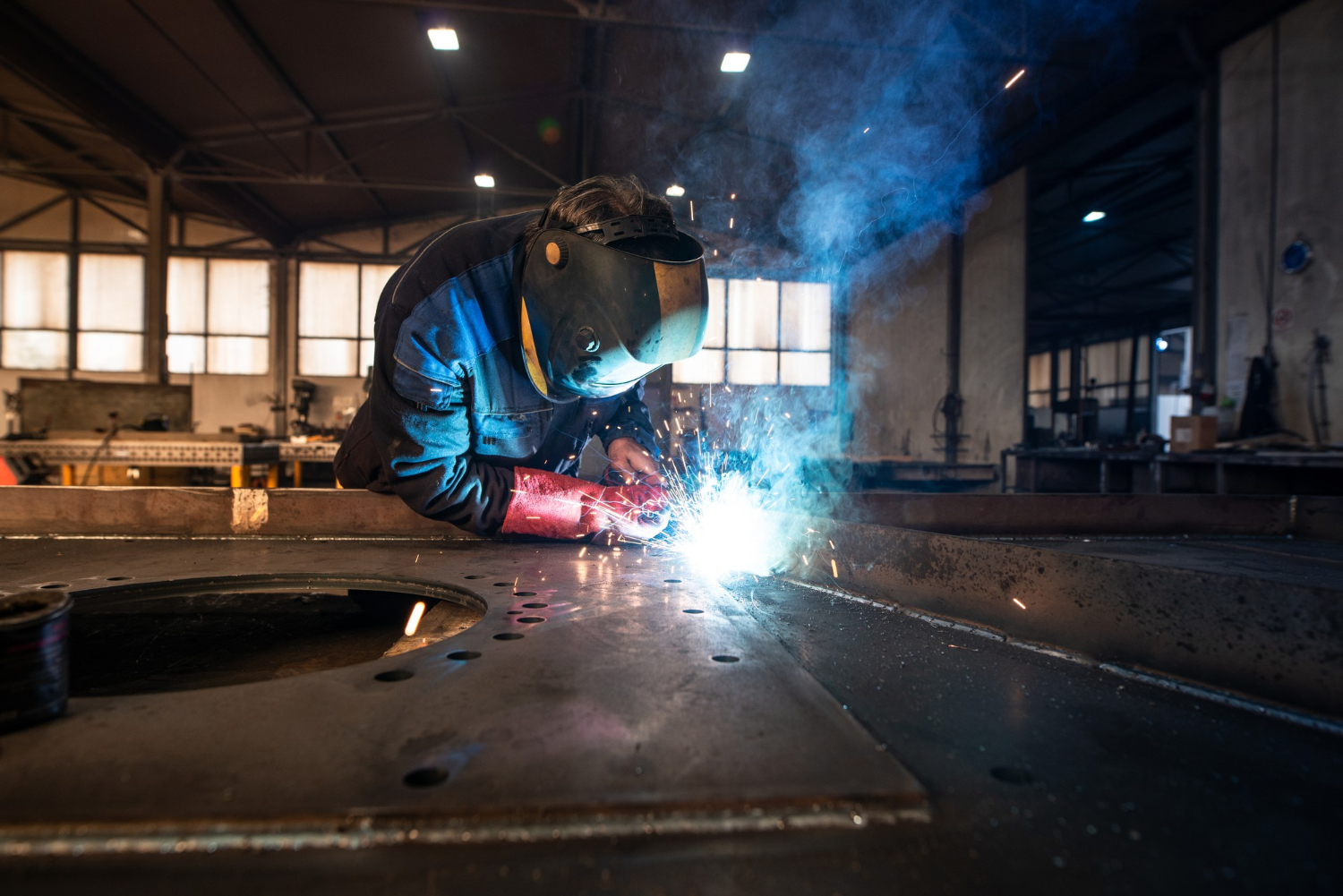 A worker in a welding mask welds metal, with bright sparks and smoke in an industrial workshop.